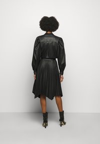 Blouse en cuir noir à manches bouffantes associée à une jupe plissée noire avec un ourlet irrégulier. Des bottines noires complètent la tenue.