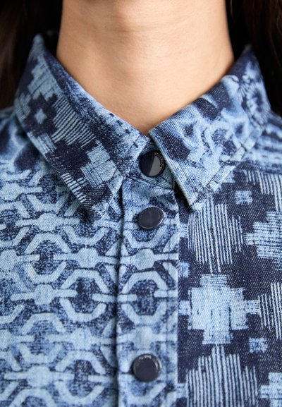 Denim shirt with a blue and navy patterned fabric, featuring a pointed collar and three black buttons. Textured surface with geometric motifs.