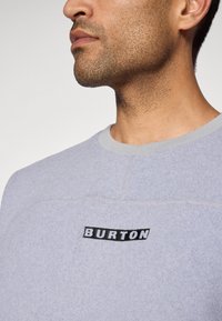Jasnoszara bluza z gładką teksturą, z czarnym logo "BURTON" na piersi oraz kontrastowym szarym ściągaczem i szwami.