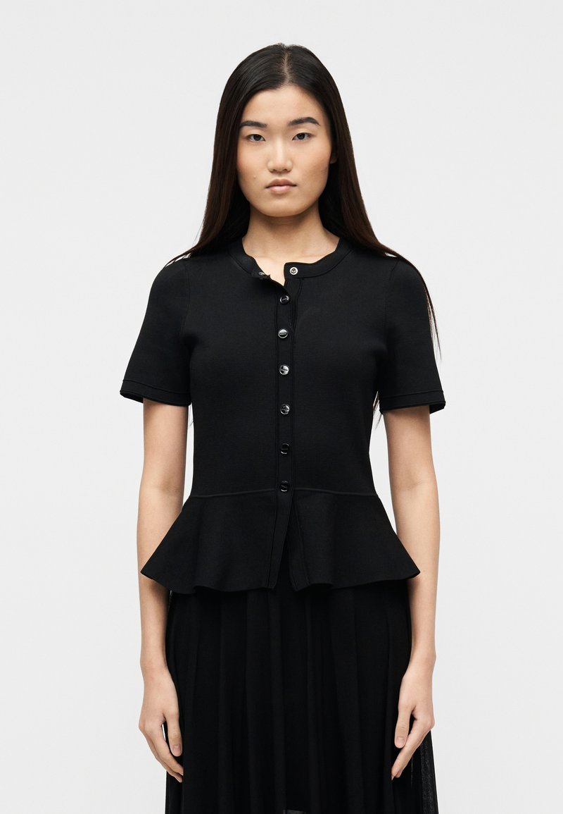 Γυναίκα με μακριά σκούρα μαλλιά, φορώντας μαύρο πουκάμισο με κοντά μανίκια και peplum τελείωμα, και μαύρη φούστα, στέκεται μπροστά σε απλό φόντο.