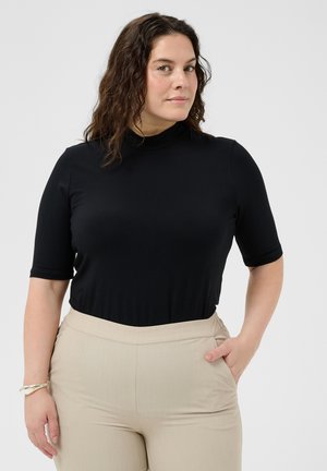 Kvinde med krøllet brunt hår iført en sort kortærmet top med høj hals og beige bukser, stående med den ene hånd i lommen.