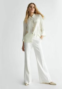 Camicia button-up gialla chiara e trasparente con colletto e taschino sul petto, abbinata a pantaloni bianchi a vita alta e gamba larga. Scarpe dorate a punta.