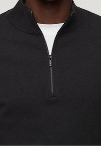 Sudadera negra con cremallera, cuello alto, tejido de punto texturizado y una cremallera corta en la parte delantera. Capa de cuello redondo blanca visible debajo.