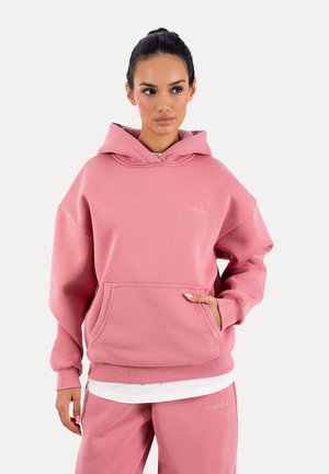 Jeune femme portant un sweat à capuche oversize rose et un pantalon de survêtement assorti, debout avec une main dans la poche avant, regardant sur le côté.