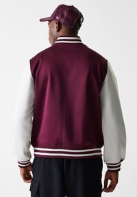 Burgunder- og hvit varsity-jakke med ribbet krage, stripete mansjetter og glatt tekstur. Kombinert med en matchende caps med nettbakside.