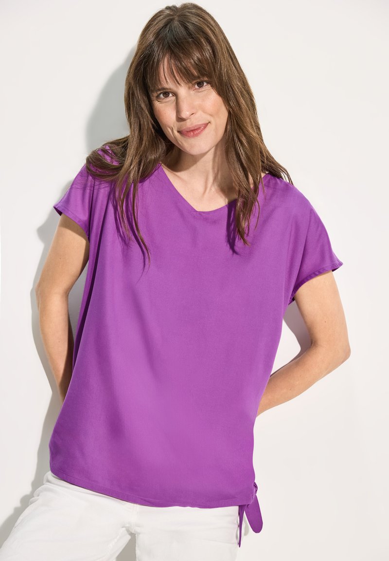 Blusa viola con maniche corte e vestibilità rilassata. Tessuto leggero, texture morbida, presenta un dettaglio a fiocco laterale per la regolazione.
