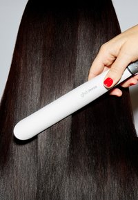 Piastra per capelli con corpo bianco e arrotondato tenuta in mano, che liscia capelli lunghi, scuri, lisci, con lucentezza visibile e texture dritta.