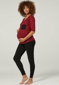 Red maternity top with a black pocket and button detail, paired with black leggings. Smooth fabric with a fitted, comfortable style.