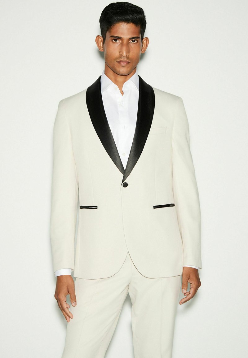 Blazer crème pour homme avec revers en satin noir, fermeture à un bouton, et accents de poche contrastants, associé à une chemise blanche.