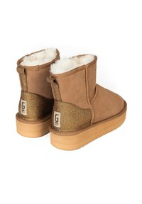 Botas de tobillo de ante marrón con un forro blanco esponjoso, detalles brillantes dorados y una suela de goma natural. Presentan una pestaña para tirar y un detalle de logo.