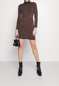Robe tricotée à motifs marron avec des manches longues et un col montant. Associée à des bottines à talons noires et à un petit sac à main noir.