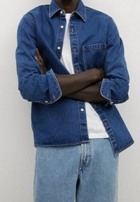 Denim overhemd in medium blauw met een knoopsluiting aan de voorkant, klassieke kraag, enkele borstzak en lange mouwen opgerold bij de manchetten. Draag het over een wit T-shirt.