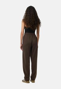 Bruine katoenen joggingbroek met een elastische tailleband; het model draagt een zwart topje en eenvoudige schoenen, gezien van de achterkant.