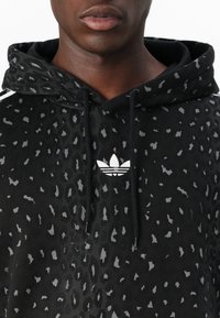 Svart hoodie med ett leopardsmönster i nyanser av grått. Inkluderar en vit Adidas-logotyp på bröstet och dragsnören i huvan.
