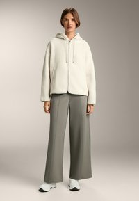 Felpa bianca fuzzy con zip e cappuccio, abbinata a pantaloni grigi loose a vita alta e sneakers bianche. Texture liscia e design casual.