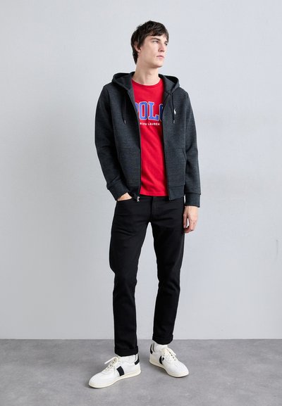 Szürke zipzáras kapucnis pulóver egy piros póló felett, kék felirattal, fekete nadrág és fehér sportcipő fekete részletekkel. Egyszerű dizájn, laza fazon.