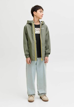 Lichtgewicht, olijfgroene regenjas met een gele ritssluiting aan de voorkant en een capuchon met trekkoord, gecombineerd met losse lichtblauwe jeans en beige schoenen.