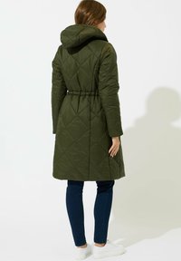 Mujer vestida con un abrigo largo acolchado de color verde oliva con capucha, jeans oscuros y zapatillas blancas, de pie con la espalda hacia la cámara.