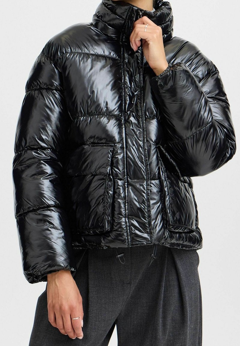 Doudoune noire avec un extérieur brillant et matelassé, col montant, fermeture éclair et poches avant. Présente un ourlet et des poignets élastiques.