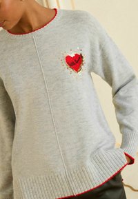 Grauer Strickpullover mit einem roten Herzmotiv und "little love", verziert mit Pailletten und roter Einfassung am Ausschnitt und Saum.