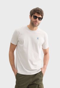Camisa de algodón beige claro con cuello redondo y mangas cortas, que presenta un pequeño logo azul con una "X" en el lado izquierdo del pecho.