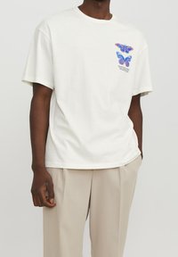 Bílý bavlněný tričko s krátkými rukávy, na prsou s grafickým motivem modrého a fialového motýla. Ležérní střih a měkká textura.