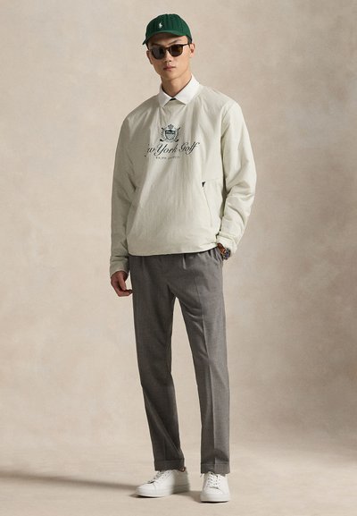 Ανοιχτό πράσινο πουλόβερ με λευκό γιακά και τύπωμα λογότυπου, συνδυασμένο με γκρι παντελόνια και λευκά αθλητικά παπούτσια. Το μοντέλο φοράει γυαλιά ηλίου και πράσινο καπέλο.