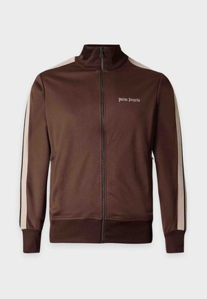 Mann steht, trägt eine braune Trainingsjacke mit cremefarbenen Streifen, ein weißes T-Shirt mit Grafik, schwarze Trainingshose mit cremefarbenen Streifen und schwarze Sneaker.