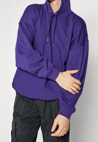 Felpa con cappuccio viola con coulisse, vestibilità oversize, tessuto morbido e polsini a coste, abbinata a pantaloni cargo scuri. Nessun dettaglio aggiuntivo visibile.