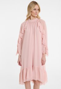 Helles rosa Kleid aus fließendem Stoff, mit Rüschendetails an den Ärmeln und dem Saum, in lockerer Passform und mit hohem Kragen.