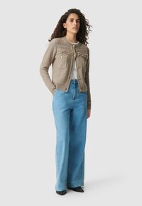 Donna con capelli ricci che indossa un cardigan di maglia beige con bottoni dorati, una maglietta bianca, jeans a gamba larga blu e scarpe nere a punta.