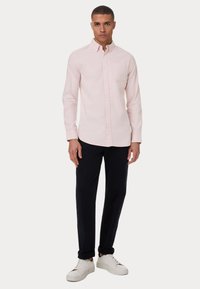 Camicia button-up rosa chiaro con maniche lunghe, taschino sul petto, abbinata a pantaloni scuri e sneakers bianche. Tessuto di consistenza liscia.