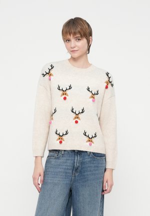 Joven vistiendo un jersey crema con caras de reno y narices de pom-pom, combinado con unos jeans azules de corte amplio, de pie contra un fondo blanco.