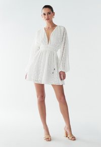 Vit miniklänning med texturerat tyg, djup V-ringning och långa ärmar. Har en elastisk midja och subtila mönstrade detaljer.