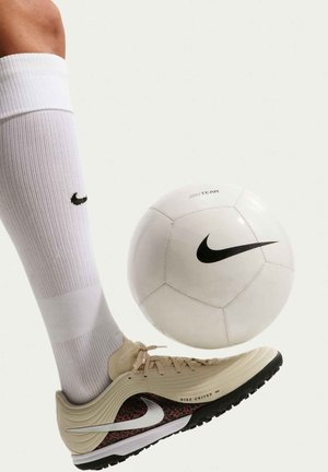 Persona con calcetín blanco Nike y zapato de fútbol beige Nike a punto de patear un balón de fútbol blanco Nike sobre un fondo claro.