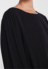 Top negro de manga larga con tejido texturizado, cuello redondo y mangas ligeramente fruncidas. Diseño minimalista sin adornos.