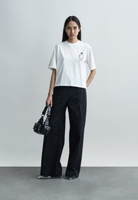 Mujer de pie contra una pared gris, vestida con una camiseta blanca con un diseño gráfico, pantalones negros de pierna ancha y zapatos plateados puntiagudos, sosteniendo un pequeño bolso negro.