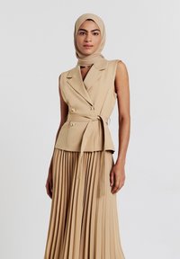 Abito beige a pieghe con top fitted senza maniche e doppio petto. Presenta una cintura in vita e bottoni dorati per un tocco in più. Tessuto dalla consistenza liscia.