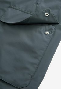 Veste imperméable vert foncé avec un rabat de poche plié maintenu par des boutons-pression argentés. Texture lisse, matériau léger, design polyvalent.