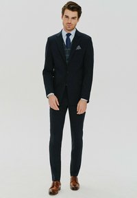 Granatowy garnitur z dopasowaną marynarką i slim fit spodniami, wzorzystą kamizelką w kratkę, białą koszulą, niebieskim krawatem i brązowymi skórzanymi butami. Chusteczka w kropki.