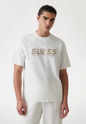 T-shirt in cotone bianco con un grande logo dorato "GUESS" e il testo "EST. 1981 - CALIFORNIA" sotto. Maniche corte, colletto a girocollo.