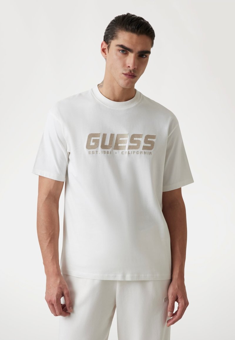 Weißes Baumwoll-T-Shirt mit einem auffälligen goldenen "GUESS"-Logo und dem Schriftzug "EST. 1981 - CALIFORNIA" darunter. Kurze Ärmel, Rundhalsausschnitt.