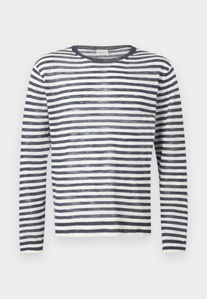 Άνδρας με κυματιστά μαλλιά που φοράει μακρυμάνικο πουλόβερ με ασπρόμαυρες ρίγες, σκούρο φαρδύ παντελόνι και μαύρα επίσημα παπούτσια, στέκεται μπροστά σε ένα απλό φόντο.