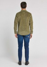 Chemise en velours côtelé vert olive avec une coupe décontractée, empiècement dos, et manches longues ; associée à un jean droit bleu foncé et des bottes noires.