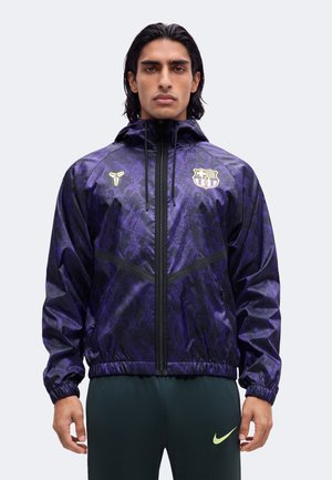 Homme portant une veste à capuche zippée violette à motifs avec le logo du club de football de Barcelone et un pantalon foncé avec un petit logo jaune Nike.