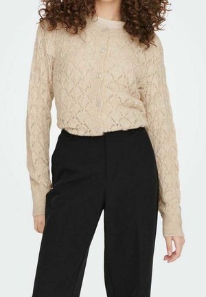 Frau trägt eine beigefarbene, geknöpfte Strickjacke mit Spitzenmuster und hochtaillierte schwarze Hose, steht vor einem einfarbigen Hintergrund.