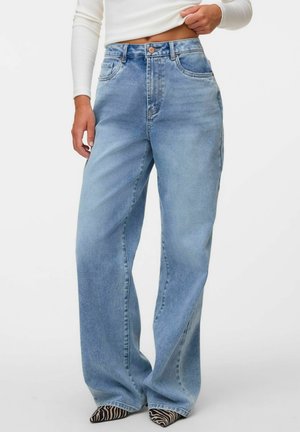 Person trägt hellblaue Jeans mit weitem Bein, weißes, kurz geschnittenes Langarmtop und spitze Schuhe mit Zebramuster vor einfarbigem Hintergrund.
