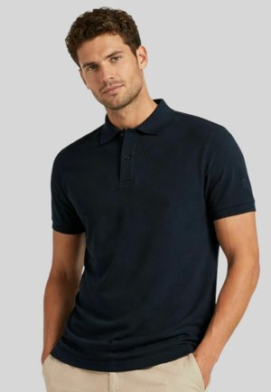 Uomo che indossa una polo blu scuro e pantaloni beige, in piedi con le mani nelle tasche davanti a uno sfondo grigio semplice.