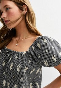 Black checkered blouse with white floral pattern, puff sleeves, and a gathered neckline. Textured fabric with a soft drape.