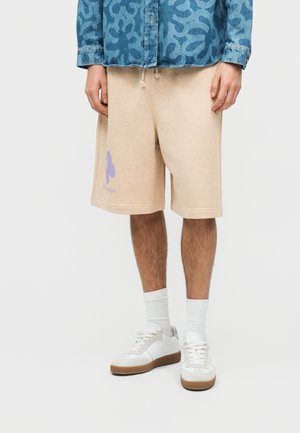 Mand iført beige knælange shorts med lilla logo på venstre ben, hvide sokker, hvide sneakers og blå mønstret langærmet skjorte.